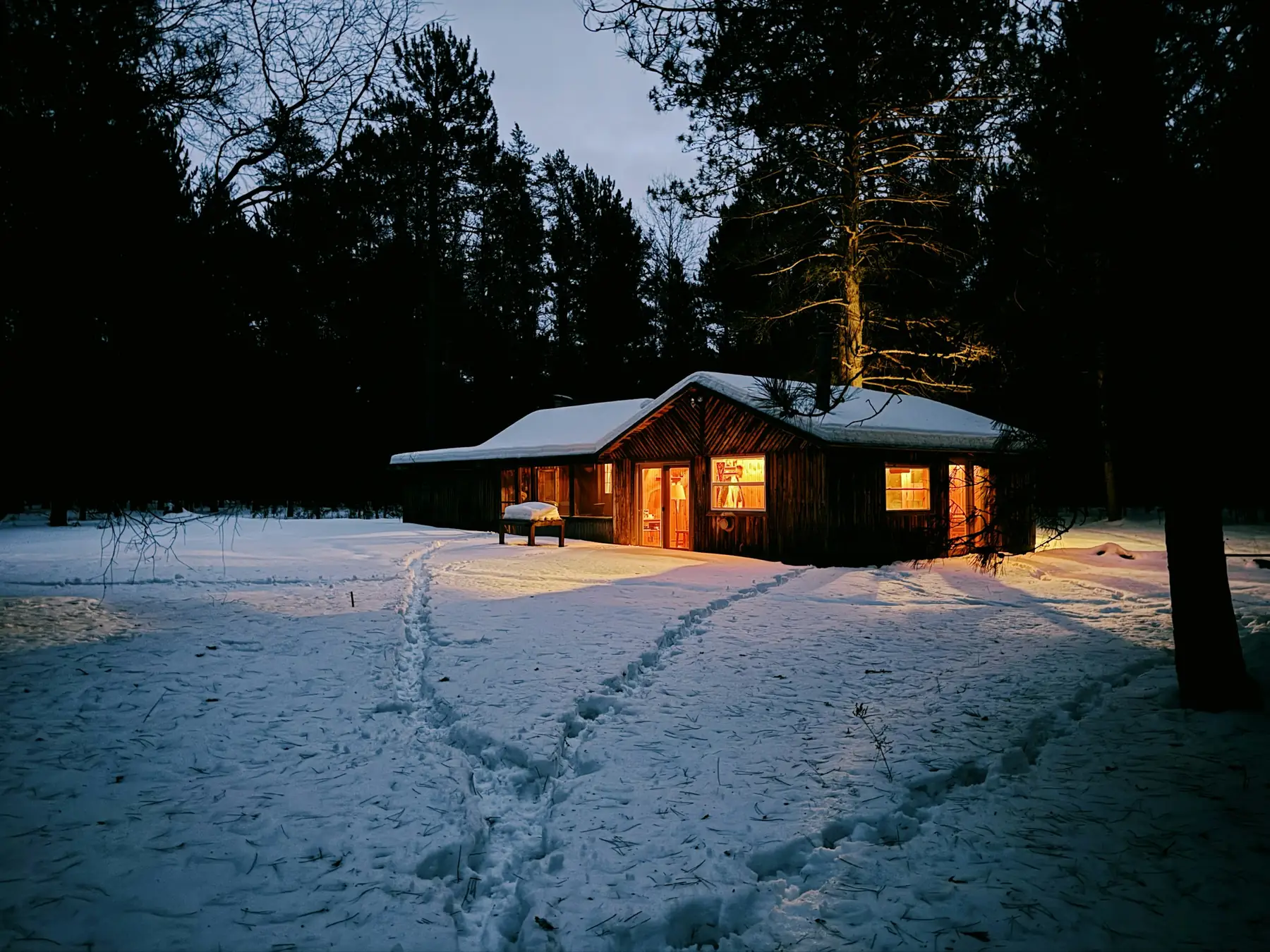 En liten trehytte i snødekt skog om kvelden, med varmt lys i vinduene.